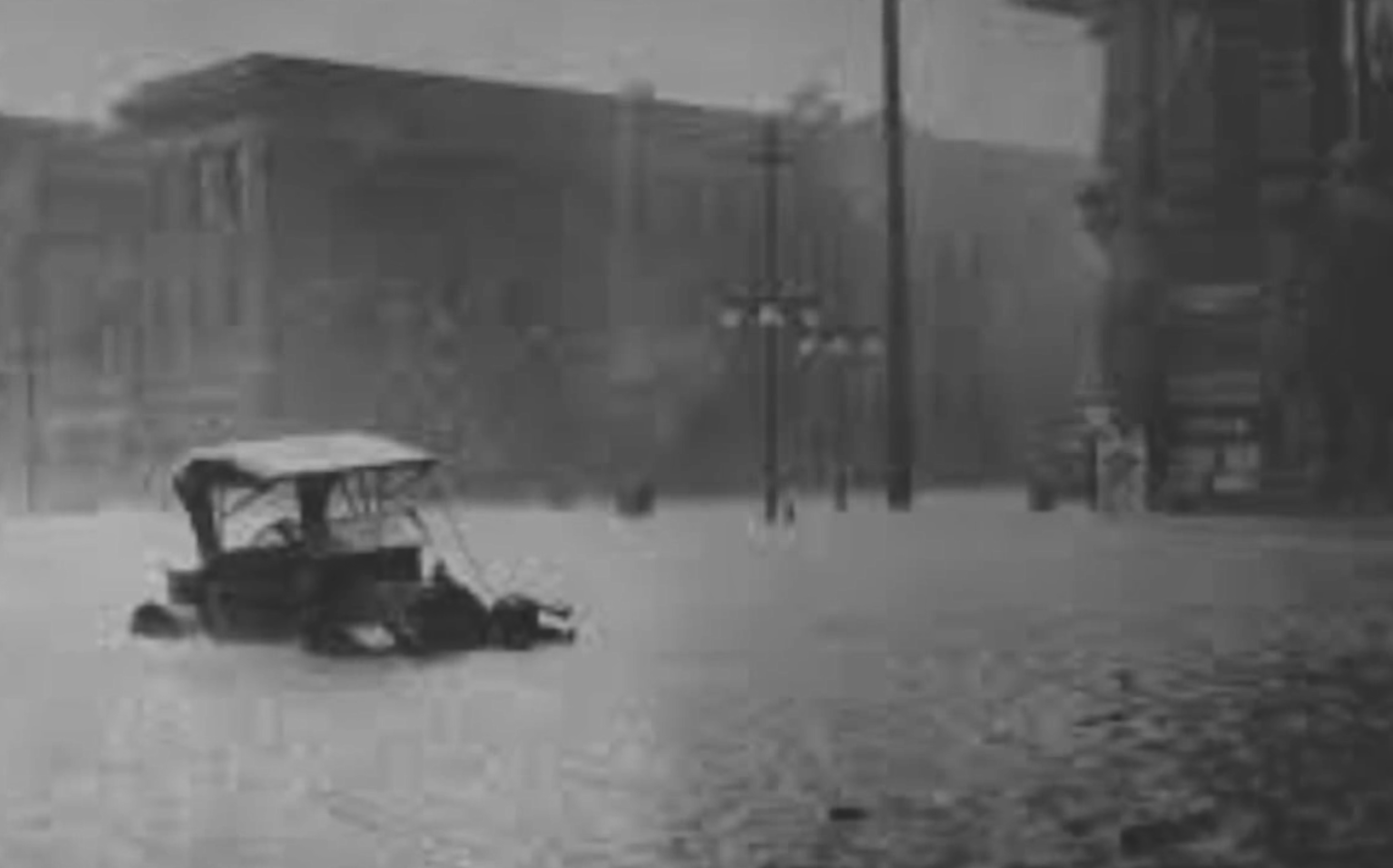 An early-20th century automobile is stuck in waste-deep water on an urban street.