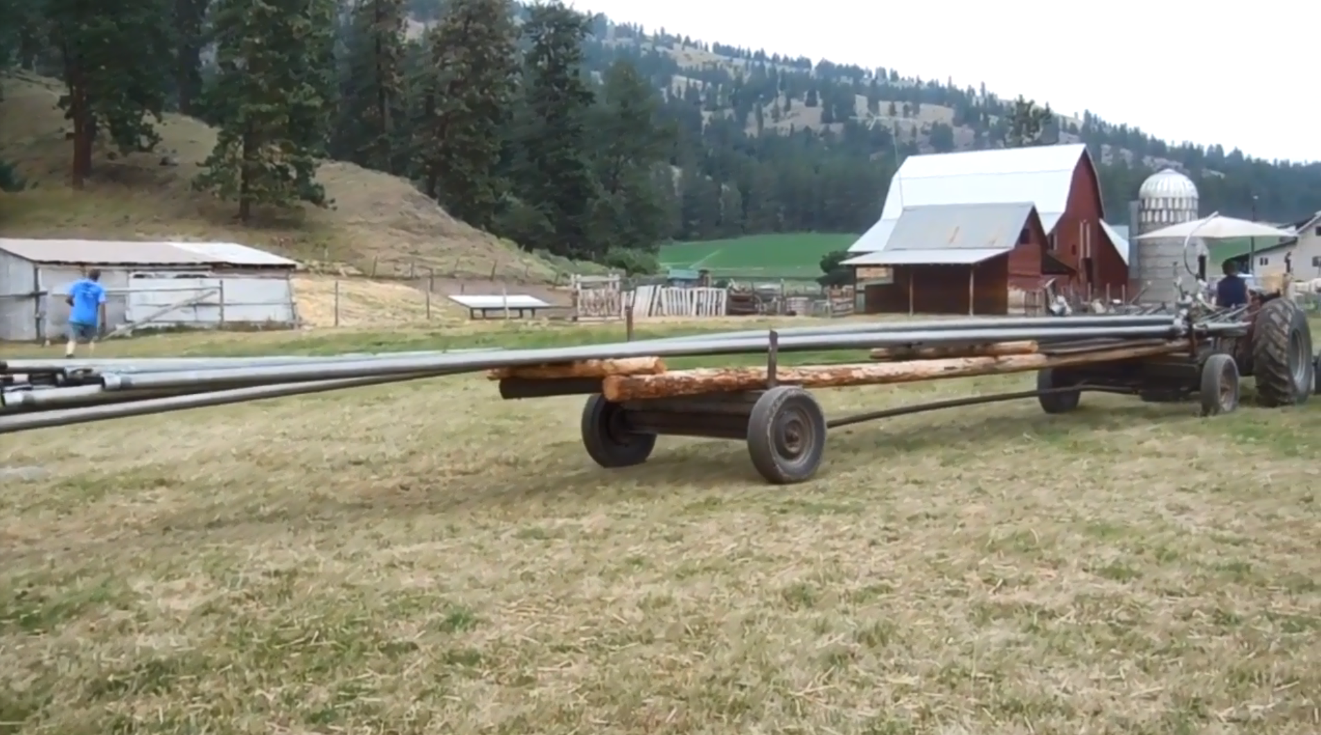 A flatbed attached to a tractor carries long pipes on the back of it as it pulls into a field on a farm.
