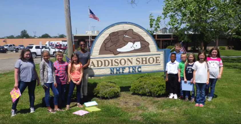 A group of students stand beside the Addison Shoe Company sign, outside the factory.