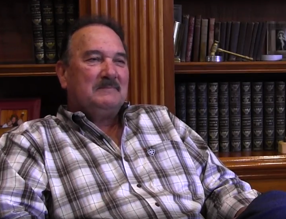 A man with a plaid shirt and a dark-colored mustache sits in a library space. 