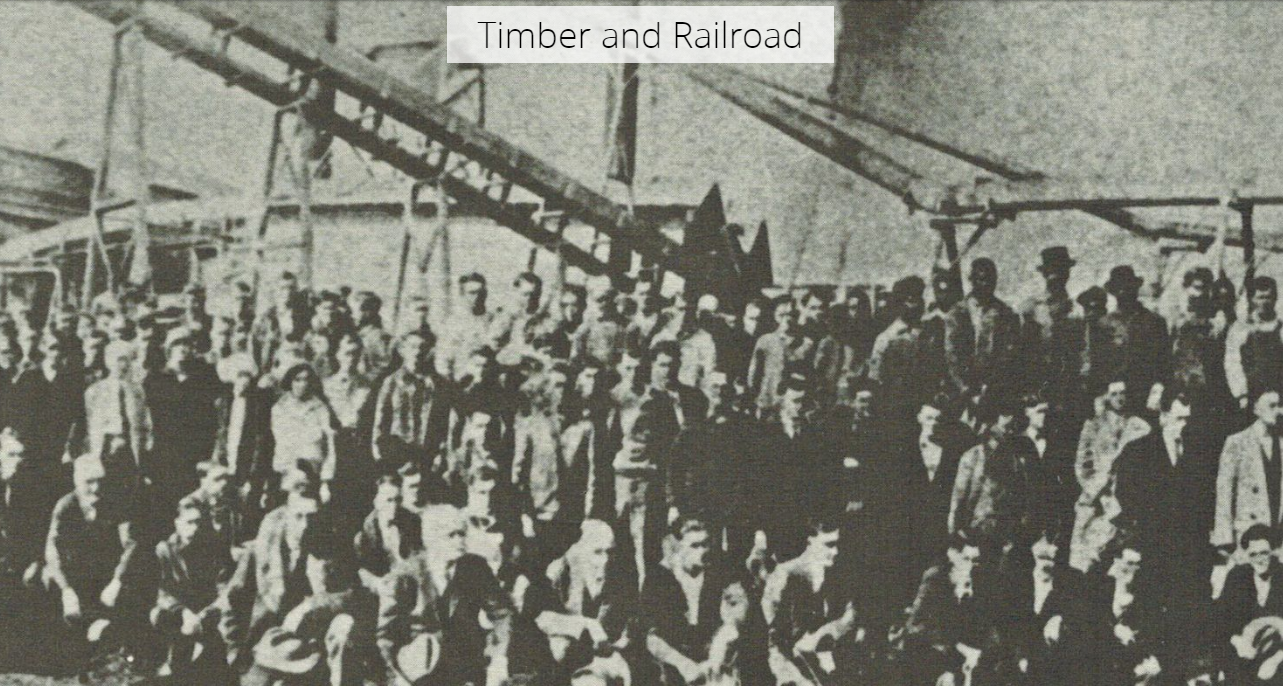 Black and white photo of a large group of men standing around machinery for timber.