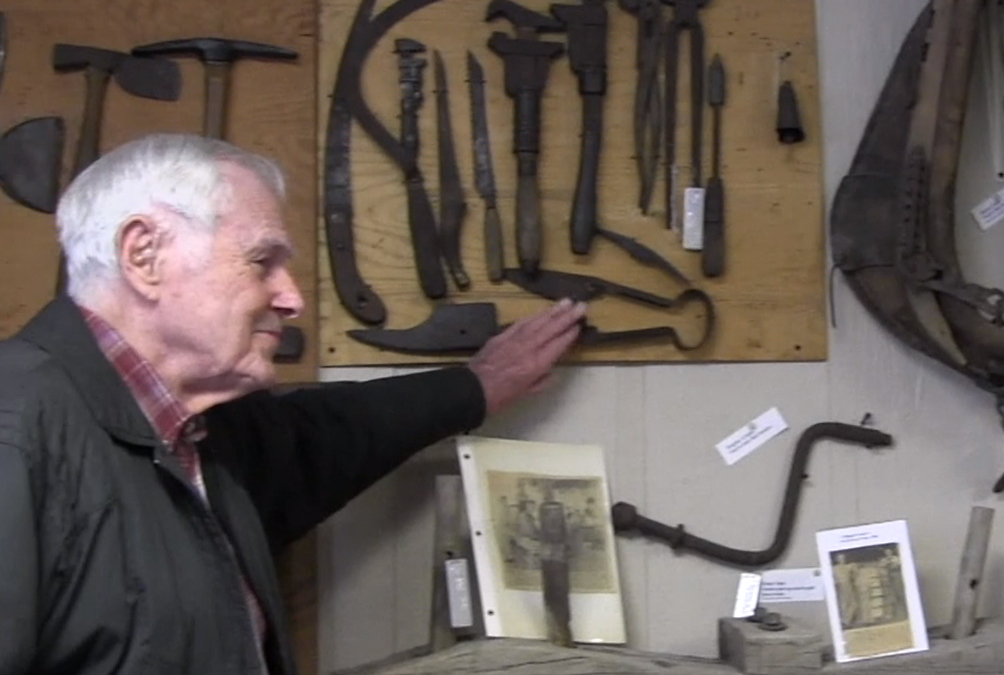 An elderly man stands next to a wall with many historic tools and points to a pair of large scissors.