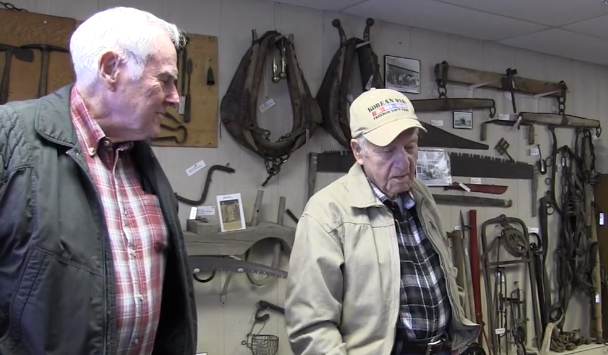 Two elderly men stand in a crowded museum space and talk about some of the objects on the walls, including old tools.