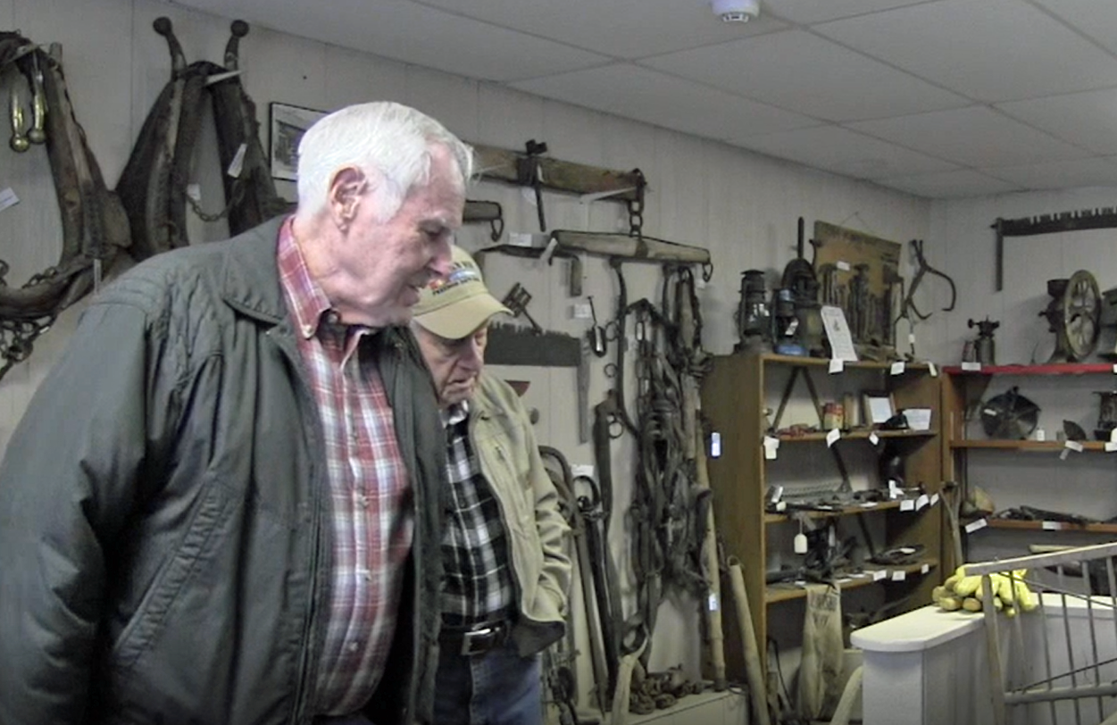 Two older gentlemen stand in a museum room filled with old farming tools.