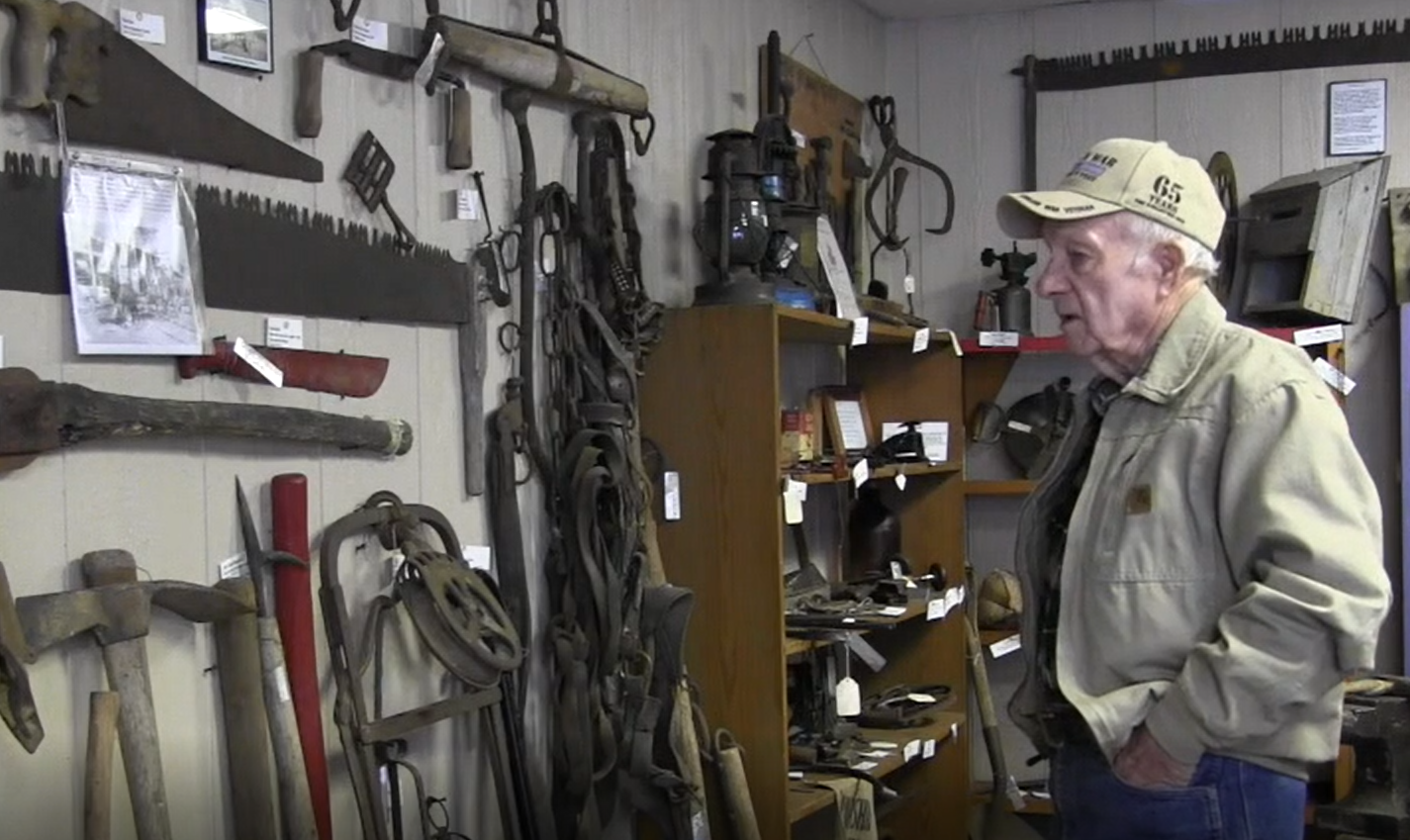 An older man in a tan ball cap and a jacket looks over a wall in a museum that displays old tools, saws, axes, and more.