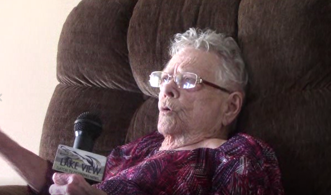 An elderly woman with gray hair and glasses sits in a plush brown chair and holds a microphone. 