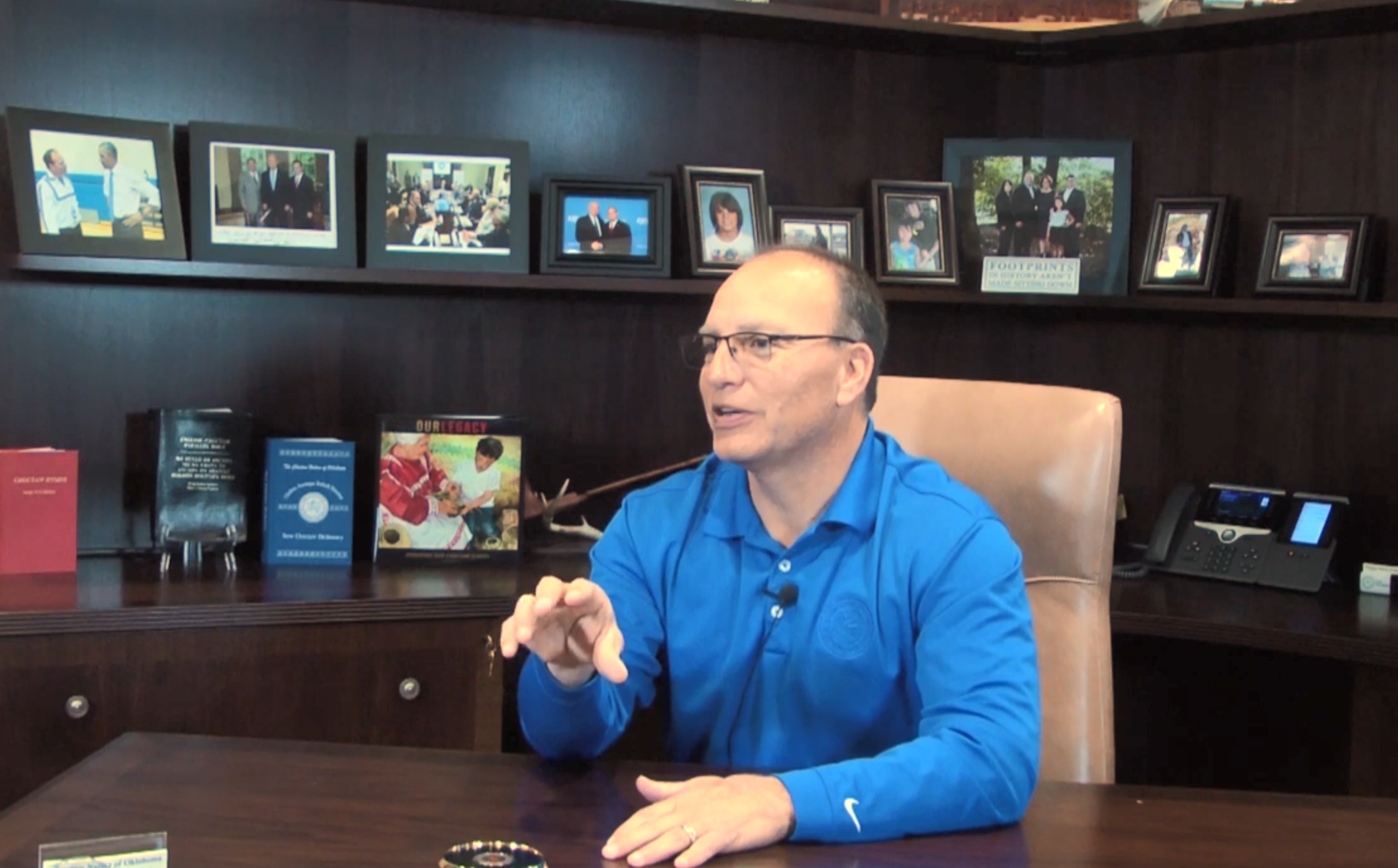 Gary wears a blue shirt and sits at his desk in an office building. 