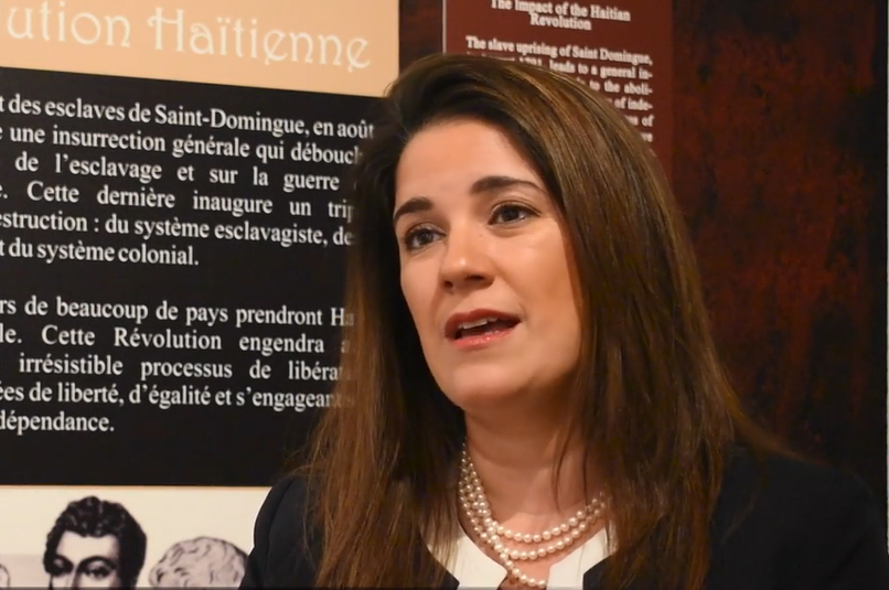 A woman with brown hair and wearing a dark jacket and pearls sits in front of a museum panel and talks to an interviewer.