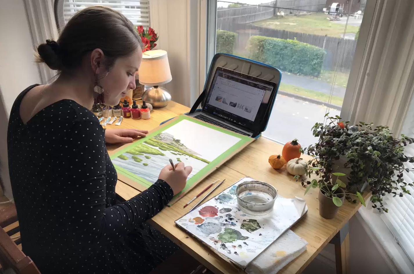 Jill sits at a desk in front of a window with an open computer, a piece of paper, and set of paints.