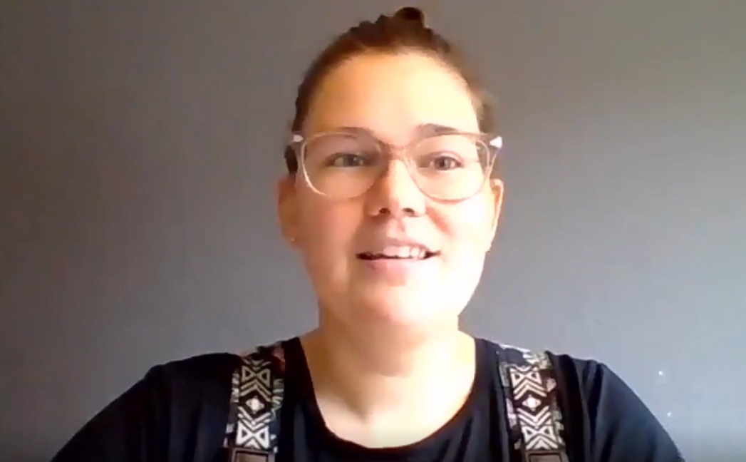 Kate wears a black shirt and clear glasses. She sits in front of a plain, dark wall.