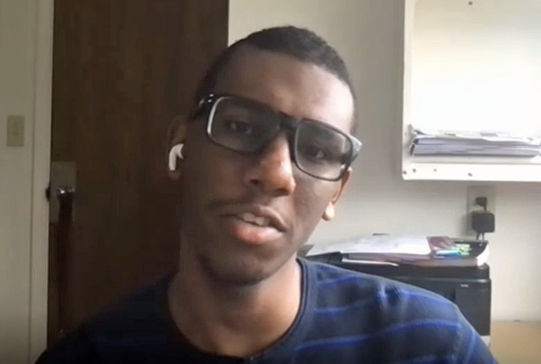 Amir has short cropped hair, a blue t-shirt, and glasses. He sits in a dorm room.