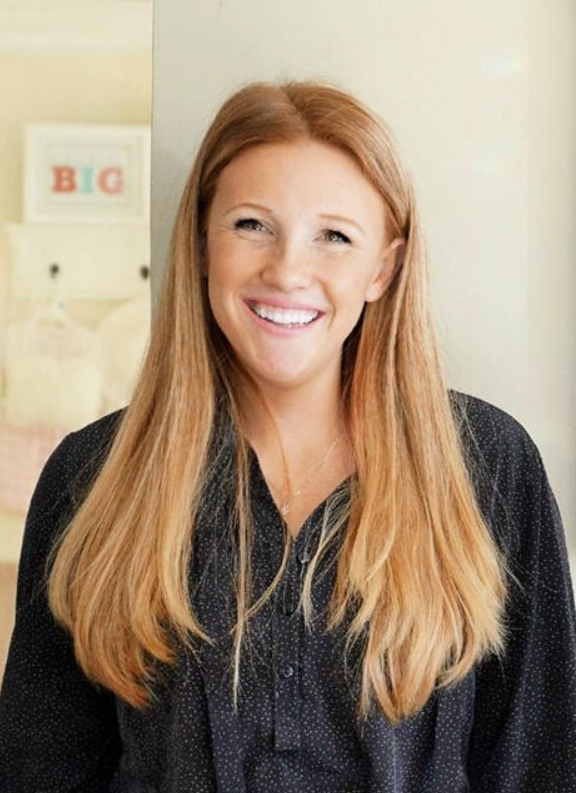 Ali stands in front of a linen-colored wall, has long, light brown hair, and a black top. 