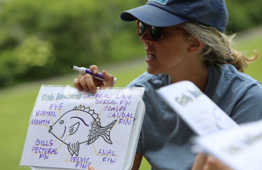 A woman with blonde hair and a blue baseball cap holds up a drawing of a fish.