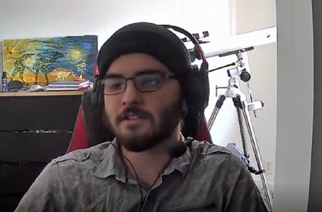 Dylan has a black knit cap on and headphones. He sits in front of a telescope and colorful painting.