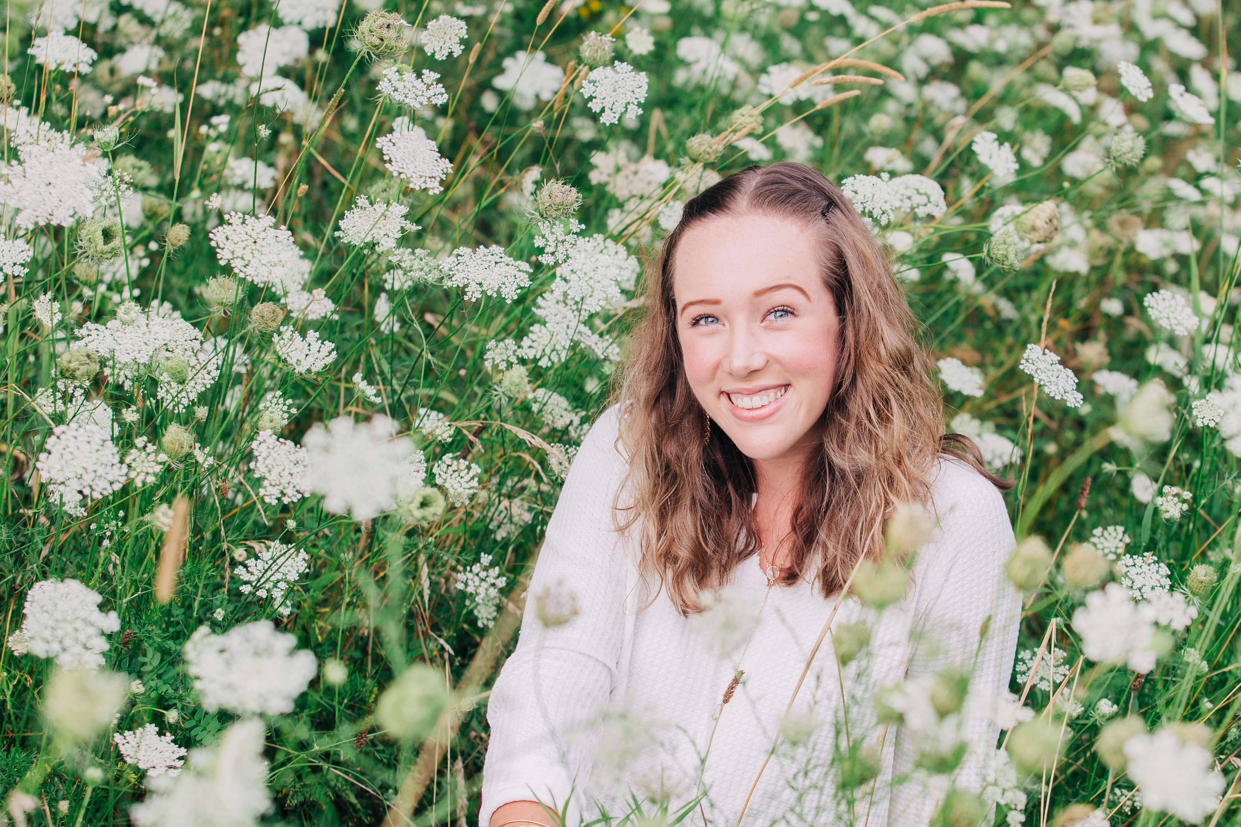 Jessica has long, brown hair and a white blouse. She sits in a field of flowers.