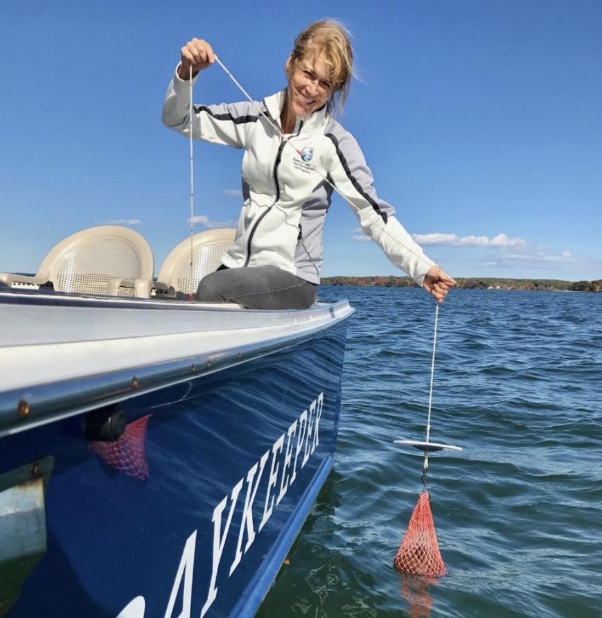 Ivy leans over the side of a boat and pulls in a line with an orange net on the bottom.