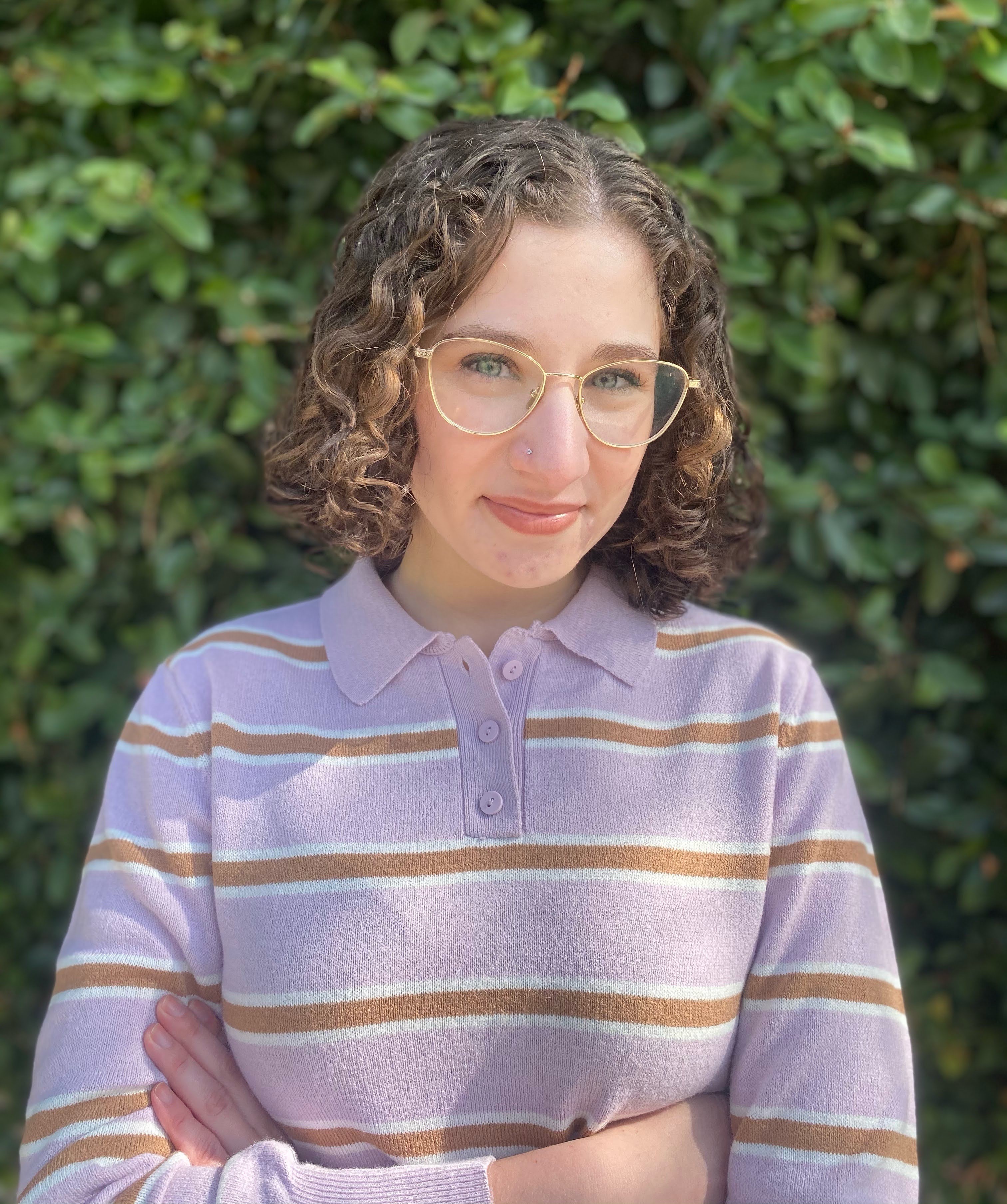 Sabrina has short, brown curly hair, eye glasses, and a purple and brown striped sweater on. She stands in front of greenery outside.