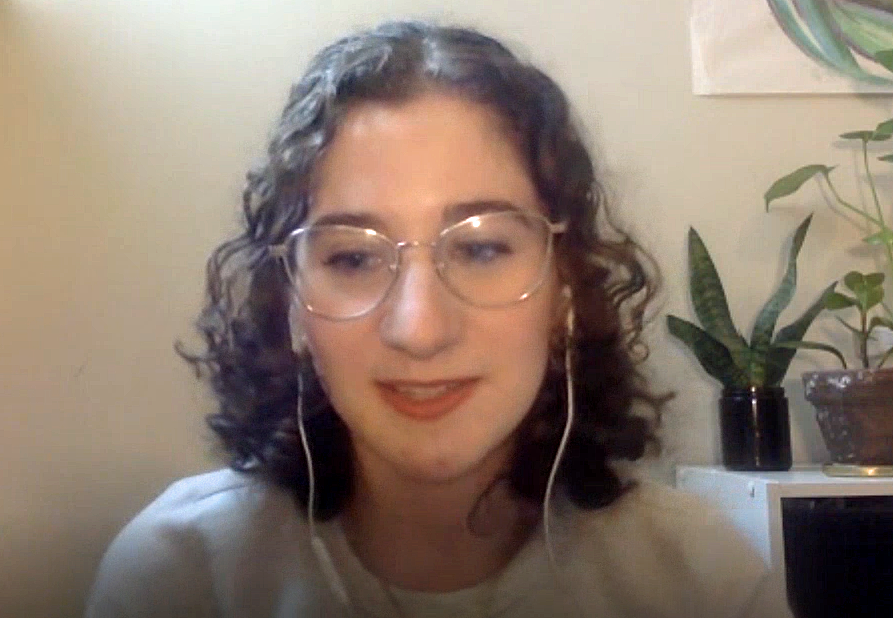 Sabrina has curly, brown hair, white headphones on, and clear-rimmed eye glasses.