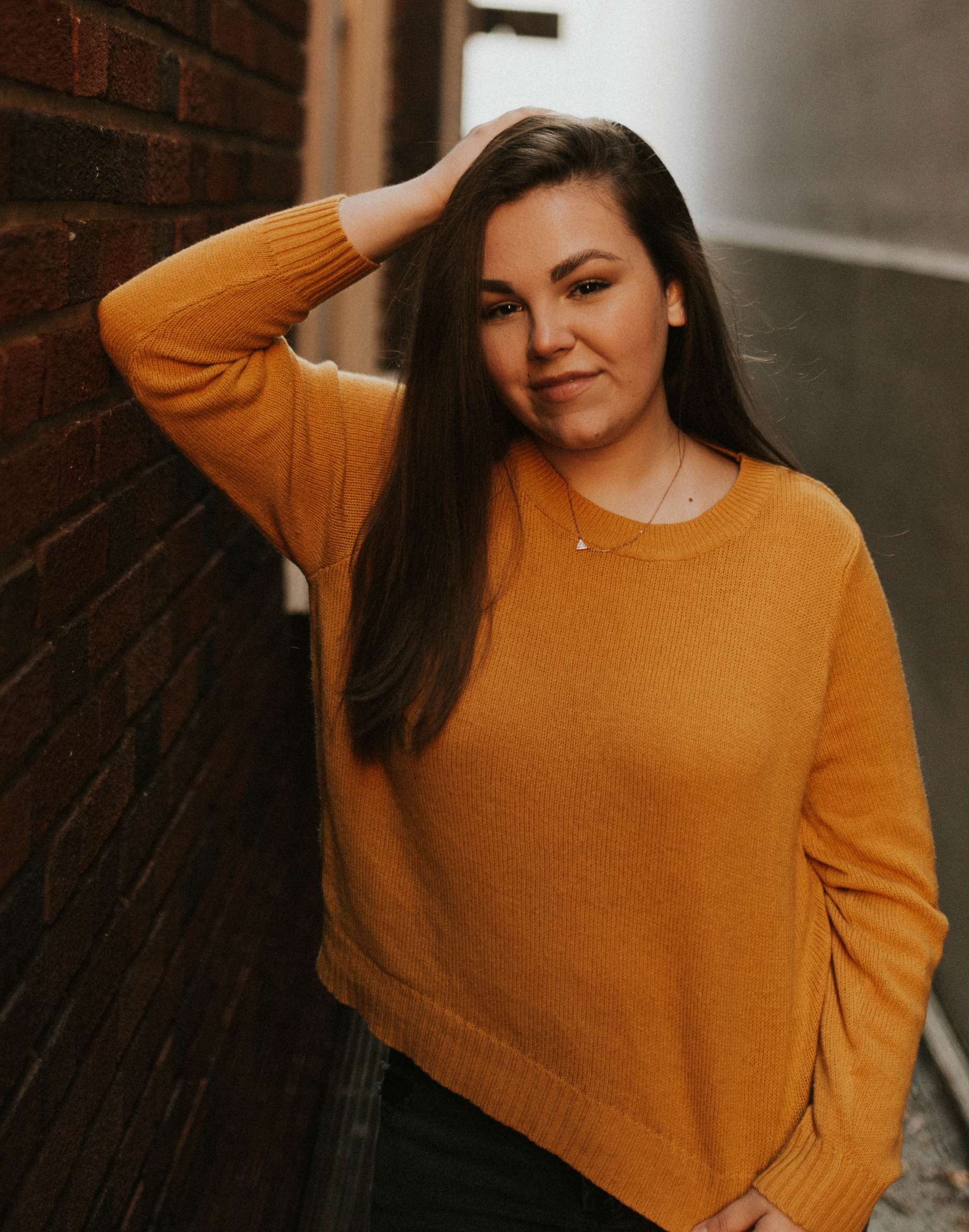 Sophia stands against the wall and puts her hand on her head. She wears a bright, orange sweater.