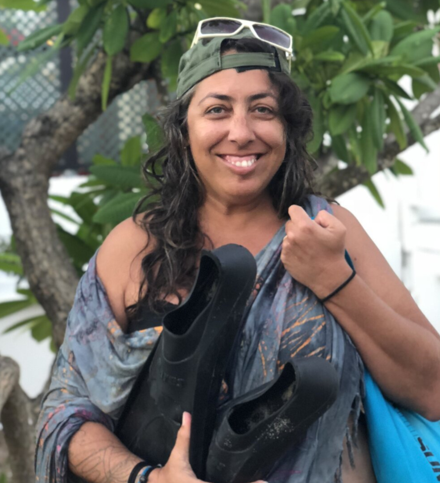 A women with sunglasses and holding flippers smiles as she stands by a tree.