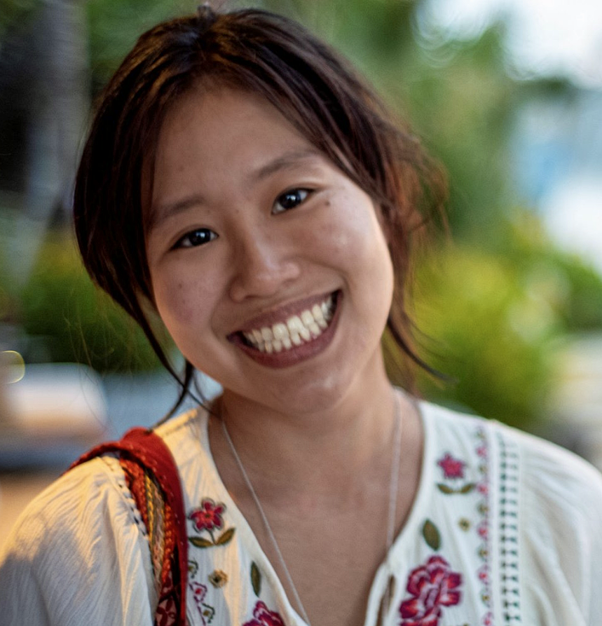 A woman with dark hair pulled back smiles broadly. She wears a top with embroidered flowers around the neck. 