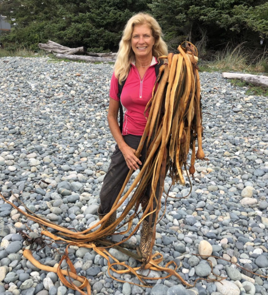A women with a hot pink shirt and long, blonde hair stands on a rocky beach and holds long strands of kelp.