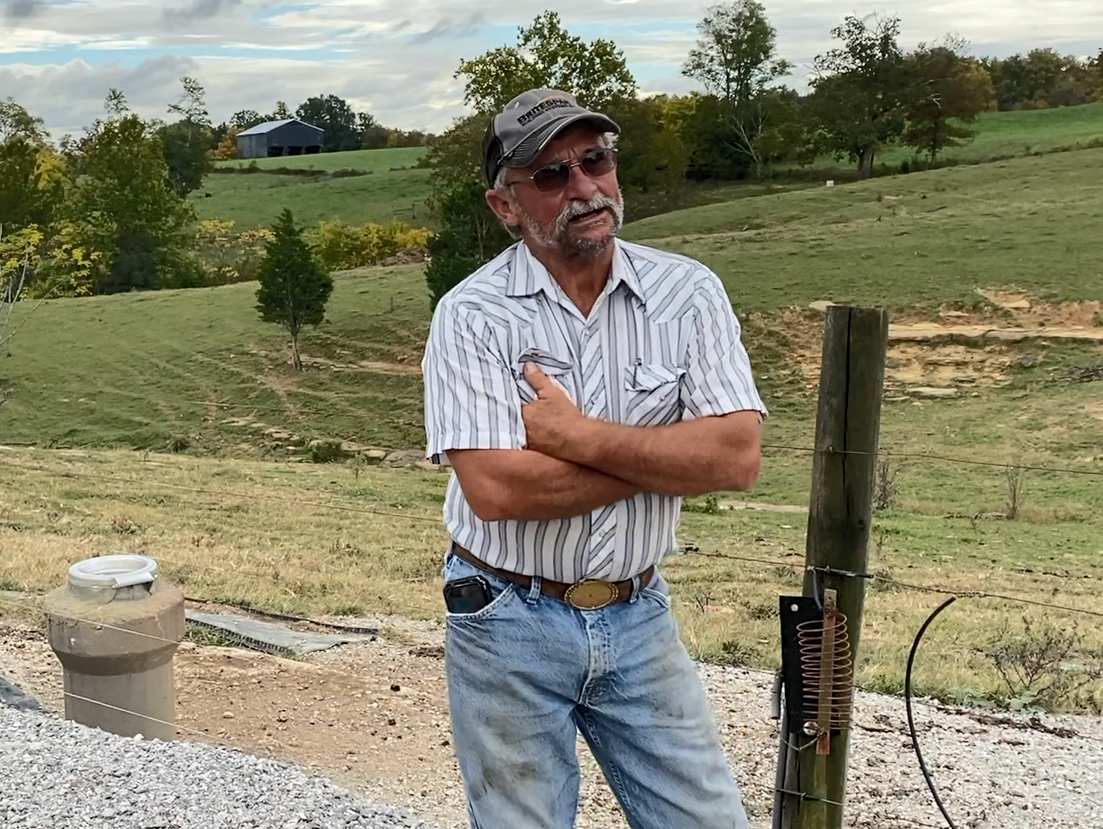 A man with a ball cap, sunglasses, and jeans stands in a farm setting. 