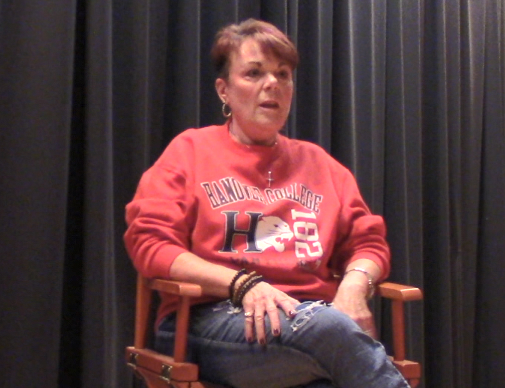 A woman with a red sweatshirt on sits in a director chair on a stage. 