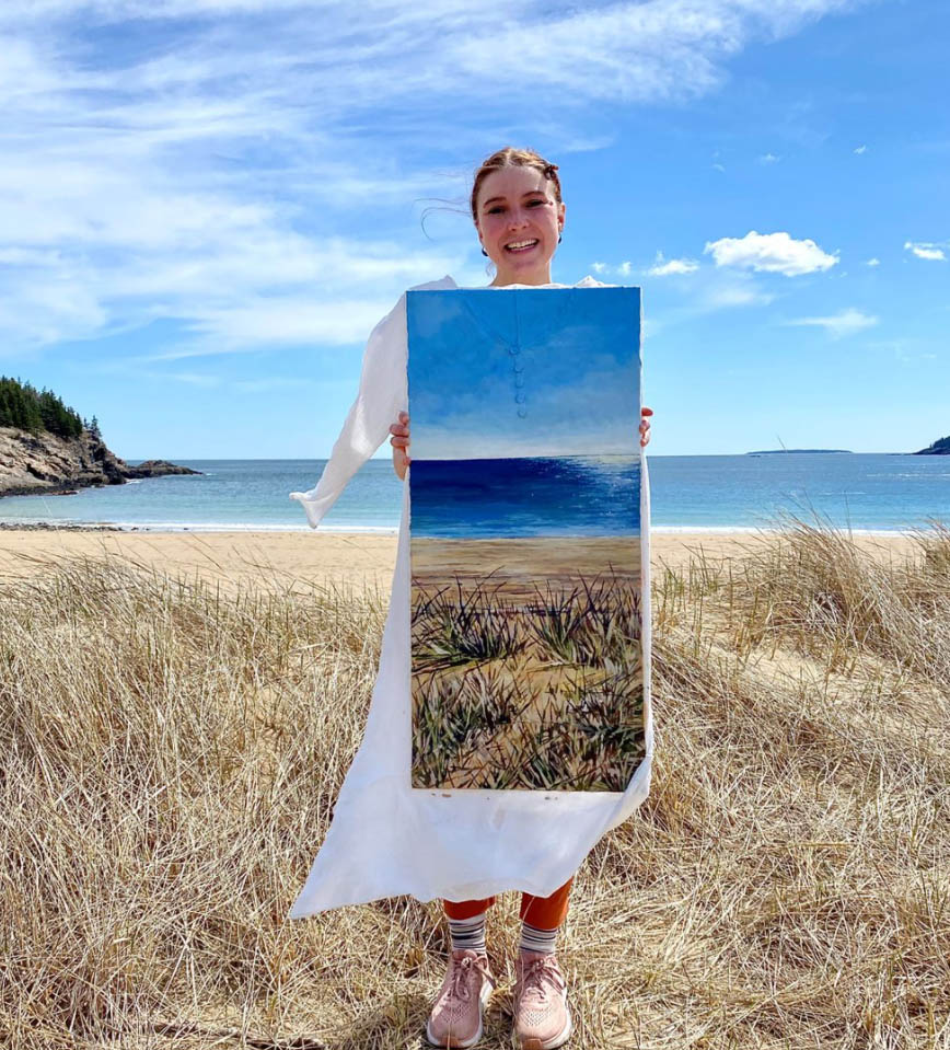 A woman stands on a beach and holds a painting up that matches the scenery exactly.