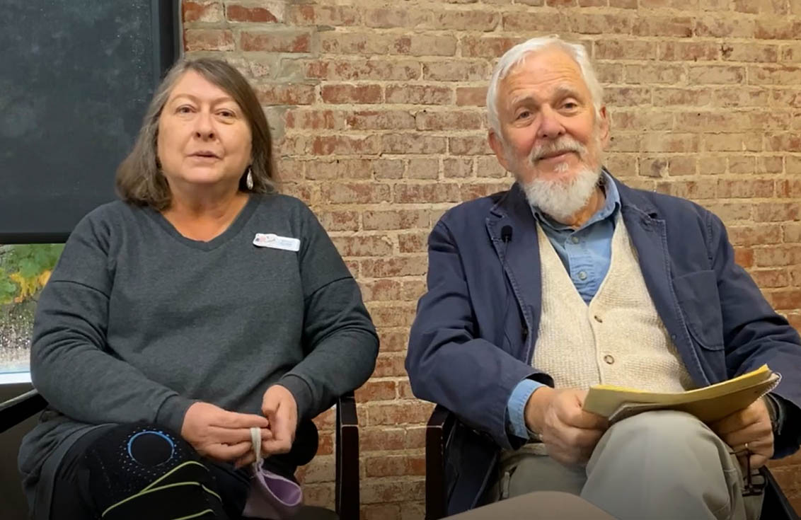 A man with white hair and a beard and a woman with shoulder-length brown hair sit in a library setting and chat with an interviewer. 