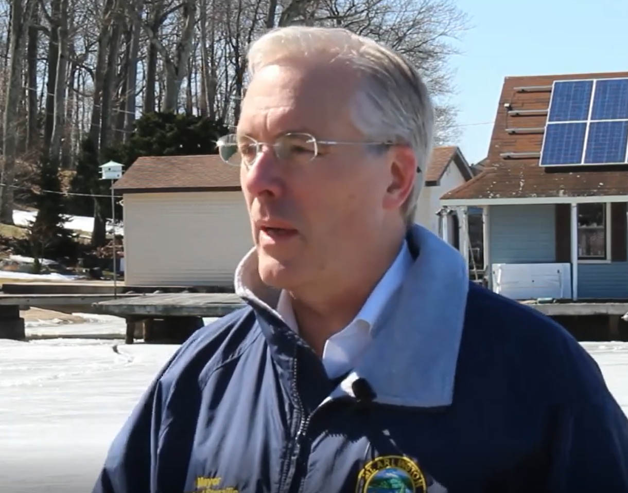 A man with white hair, glasses, and a blue winter jacket stands outside in a parking lot and chats with an interviewer.