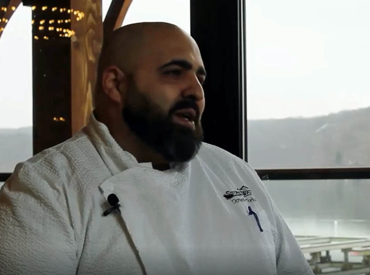 A man with a bald head and a thick brown beard wears a chef's jacket and sits in front of a lake.