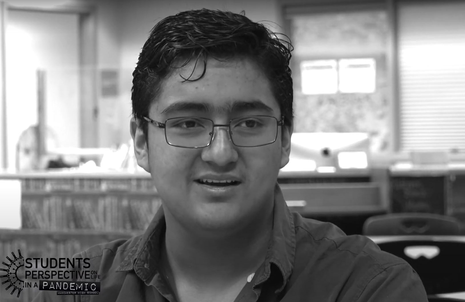A young man with short, black hair and wire frame glasses sits in a library setting and is interviewed.