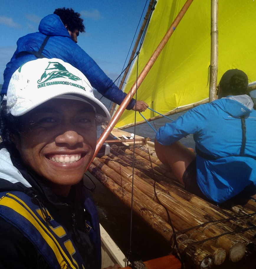 Suzanne wears a white baseball cap and sits on an outrigger on the water with a bright yellow sail.