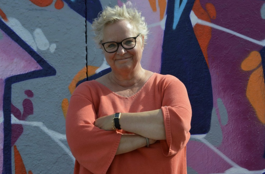 A woman with black-rimmed eye glasses and spikey blonde hair stands in front of a colorful, abstract wall mural. 