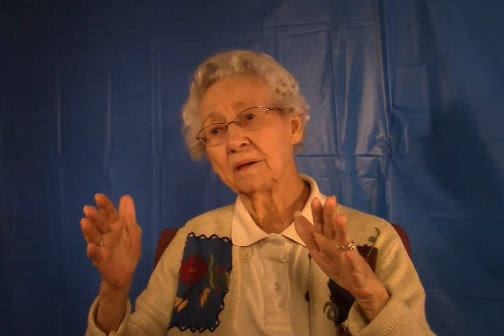 A woman with white hair and colorful vest sits in front of a blue backdrop.