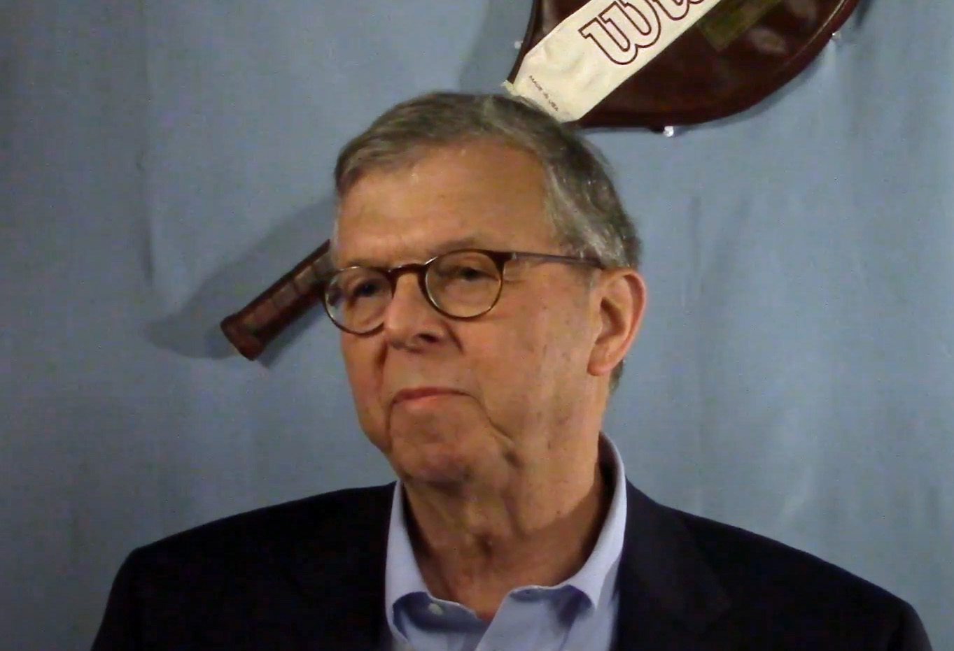 A man with glasses and blue sport jacket sits in front of a blue curtain and a tennis racket.