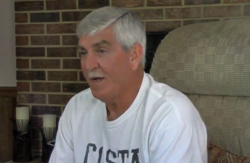 A man with white hair sits in a brown arm chair by a brick fireplace. 