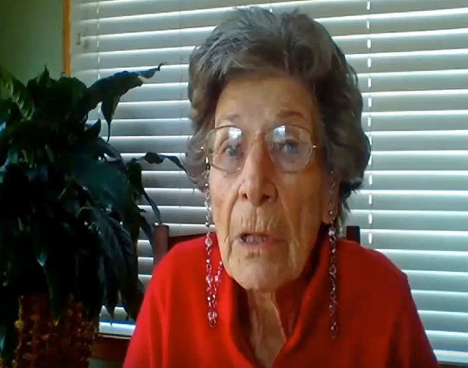 Mrs. Griffin wears a bright red blouse and eyeglasses. She sits in front of a window with blinds and a plant.