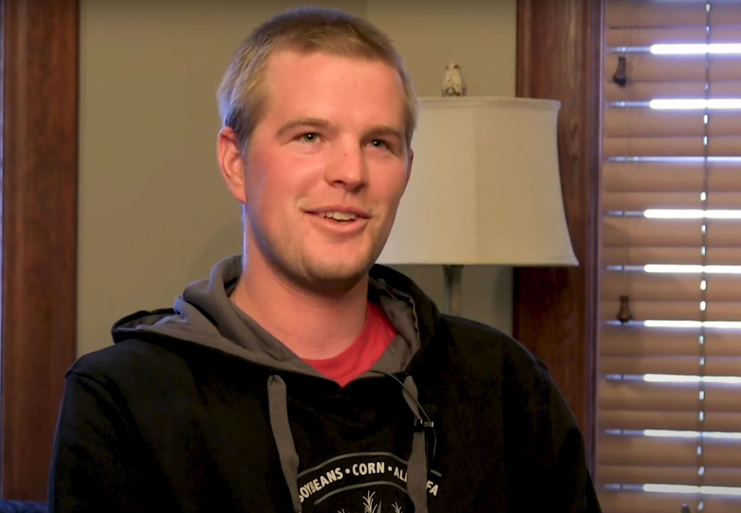 A young man with short blonde hair and a black sweatshirt sits on a couch with a window behind him.