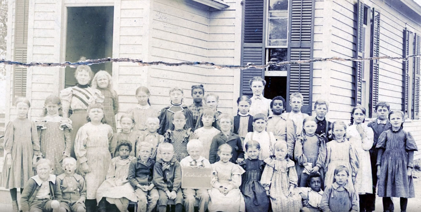 A large group of children stand in front of an old school house around the turn of the 20th century.