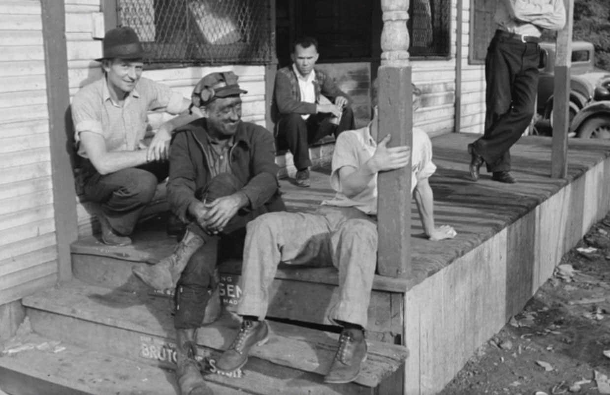 A 1930s image of men sitting on the wooden front porch of a house. 