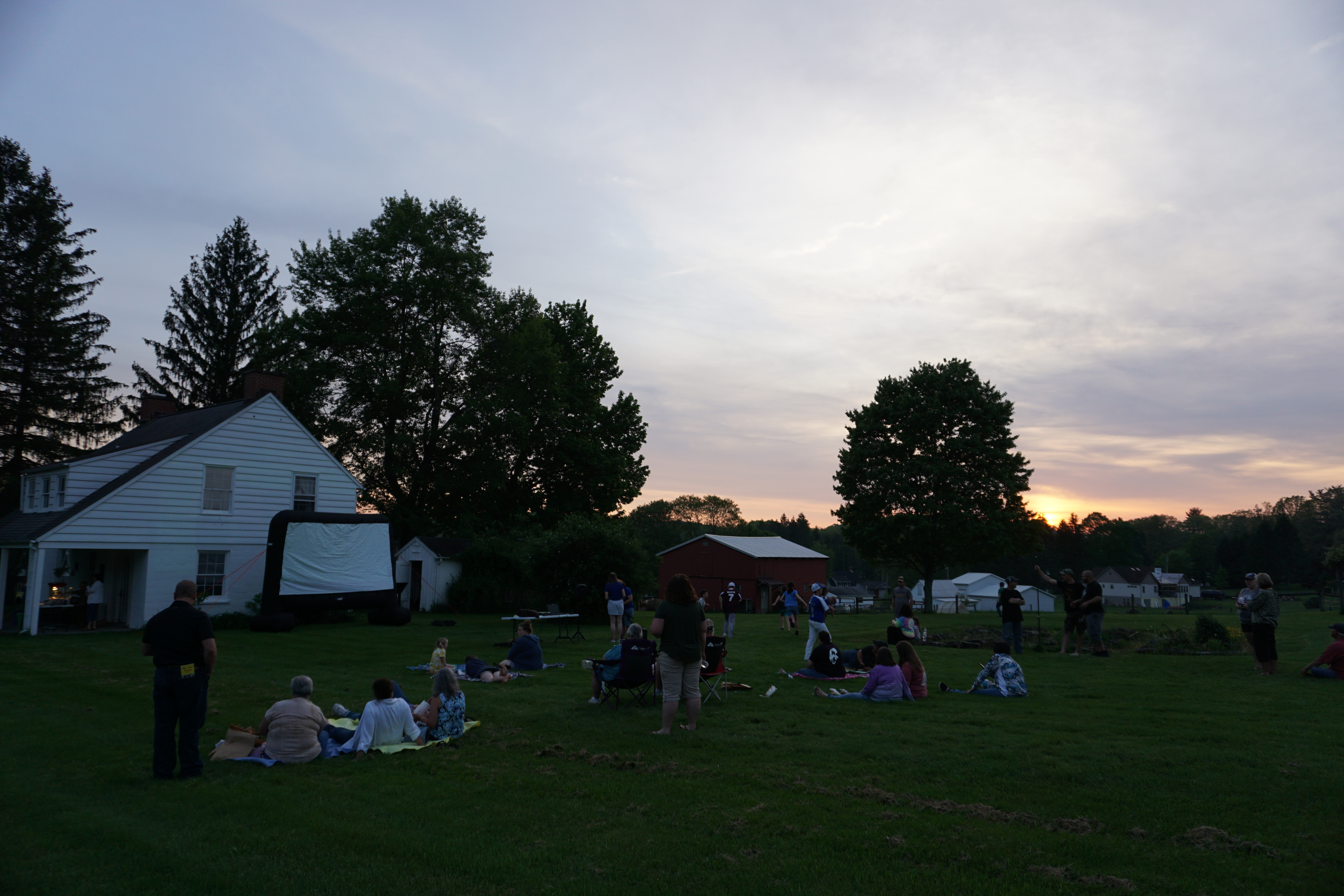 A white farm building and people gathered on the lawn of it at dusk.