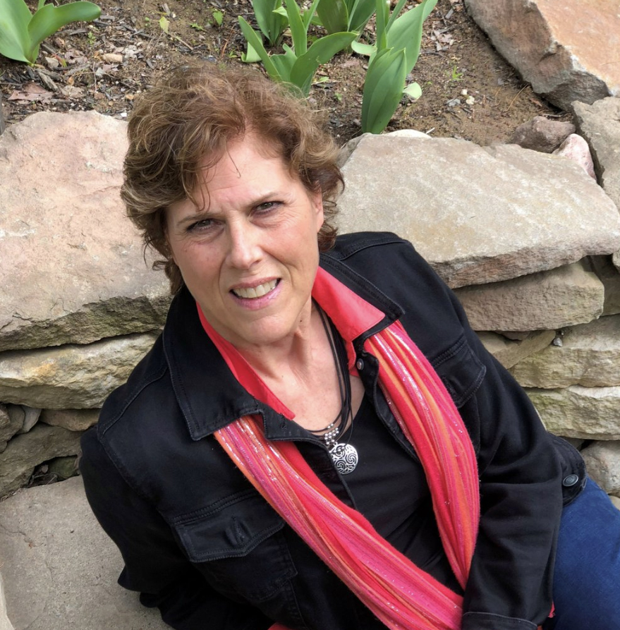 A woman with brown hair and a bright pink scarf sits on a rock ledge. 