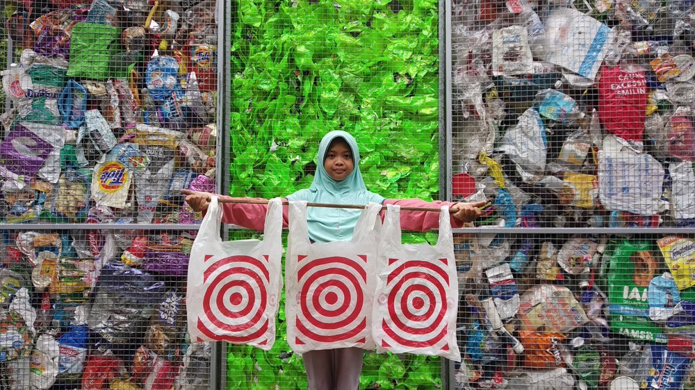 A girl stands in front of a wall of plastic trash. She's holding Target plastic bags.