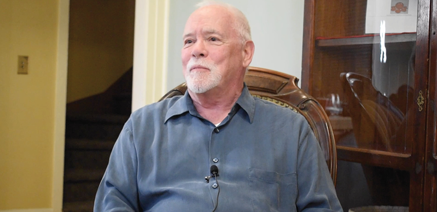 A man with a white beard wears a button-down blue shirt and sits in a vintage chair for an interview.