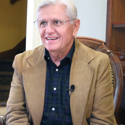 A man with white hair and a suede brown jacket sits in a vintage chair. 