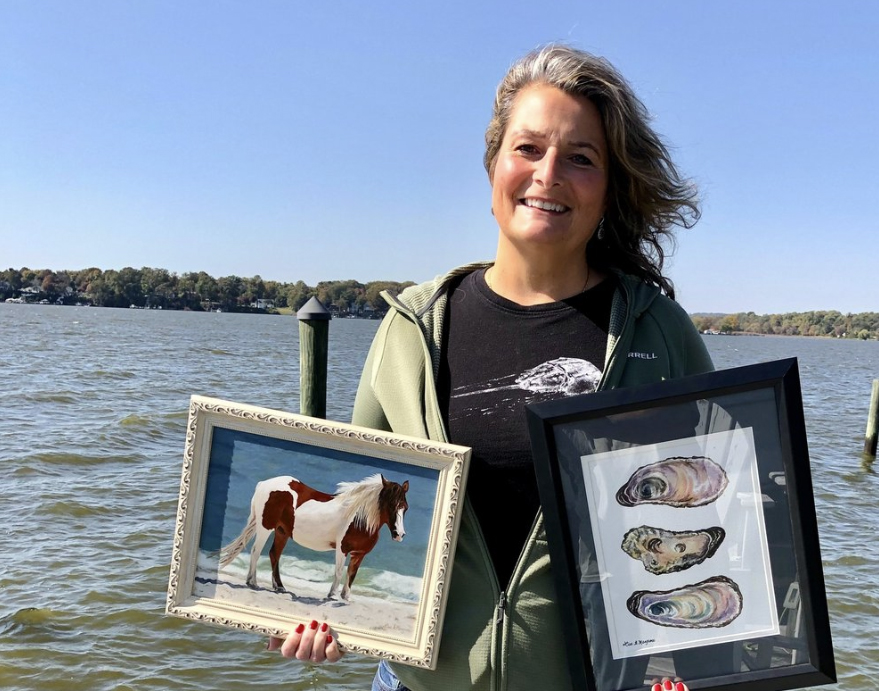 A woman stands on a dock by the water and holds paintings in her hands.