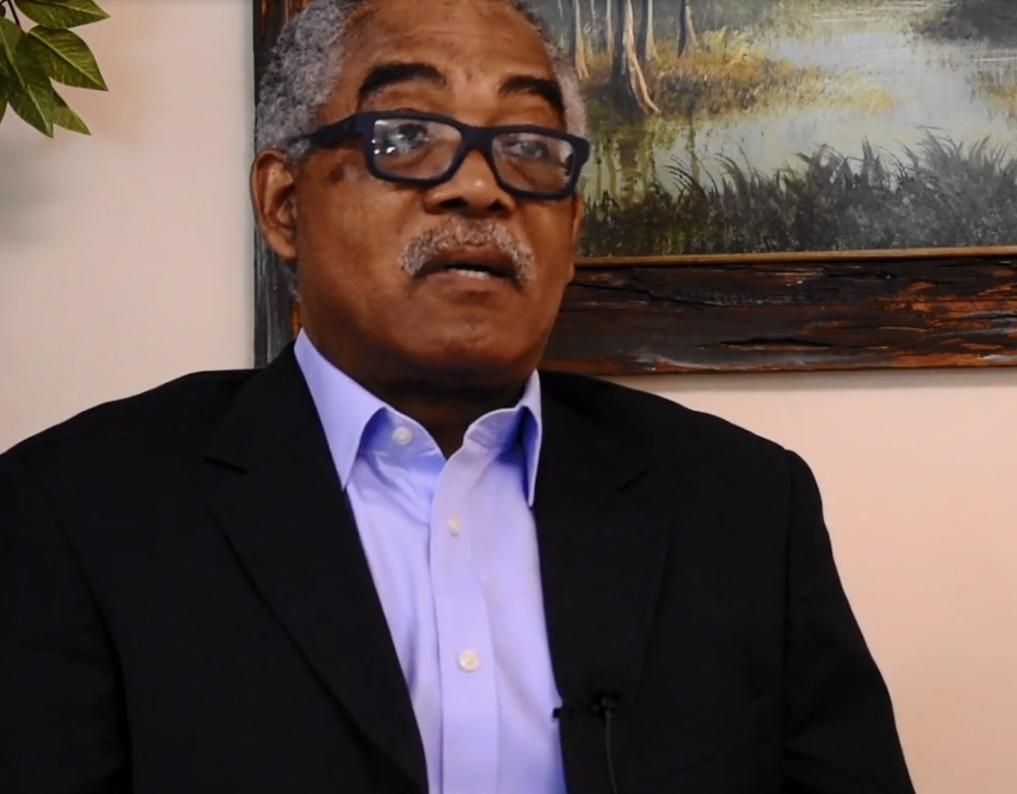 A man with gray hair, black glasses, and a purple button-downed shirt sits for an interview. 