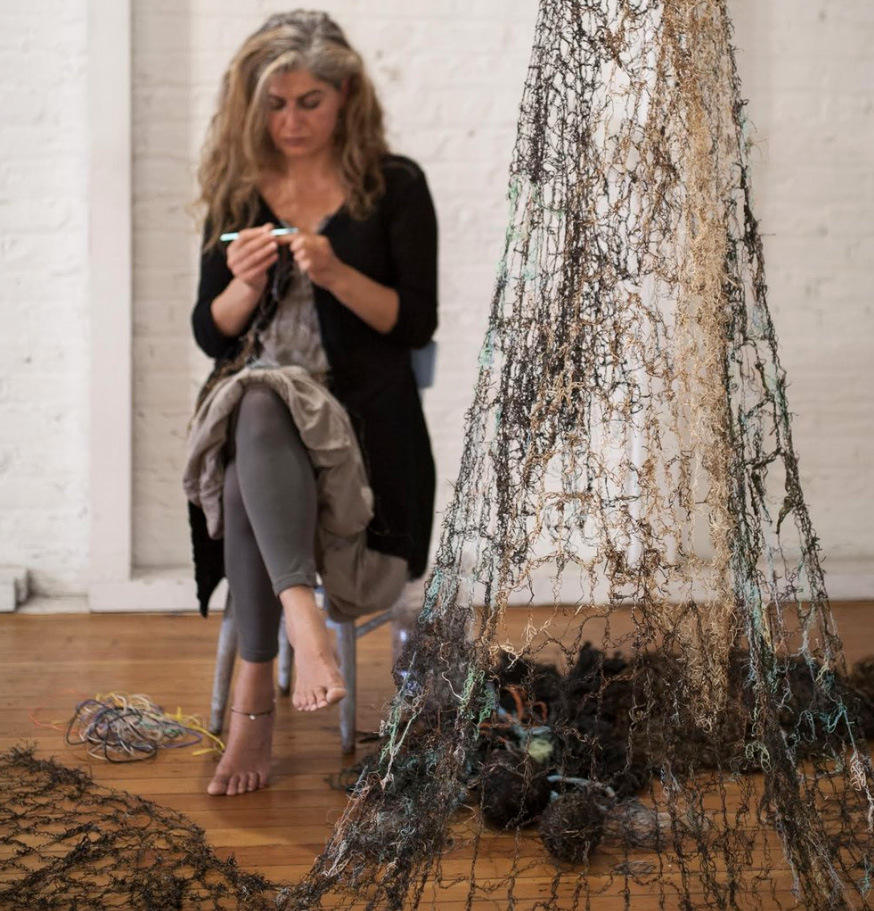 A woman with long hair and a black sweater sits and crochets marine debris into netting. 
